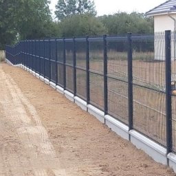 SafeFence Ogrodzenia - Długie, czarne ogrodzenie panelowe z podmurówką betonową, ciągnące się wzdłuż piaszczystej ścieżki, z domem w tle. Widok z perspektywy bocznej.