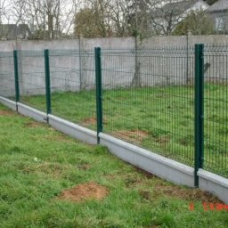 SafeFence Ogrodzenia - Nowoczesne ogrodzenie panelowe z zielonymi słupkami i betonową podmurówką na trawniku, widok z boku, w tle szary mur i domy.