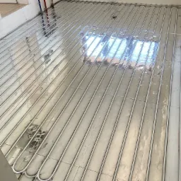 Układanie rur ogrzewania podłogowego na płytkach, widoczne złącza instalacji wodnej przy ścianie, surowy stan wykończenia.