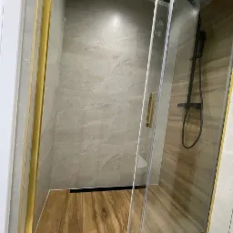 Kabina prysznicowa z przesuwnymi drzwiami, złote ramy, drewnopodobna podłoga, czarna armatura, szare płytki na ścianach.