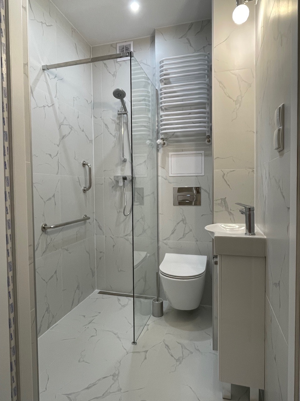 Nowoczesna łazienka w bieli i szarości z kabiną prysznicową typu walk-in, umywalką z szafką i wiszącą toaletą, wykończona płytkami imitującymi marmur.