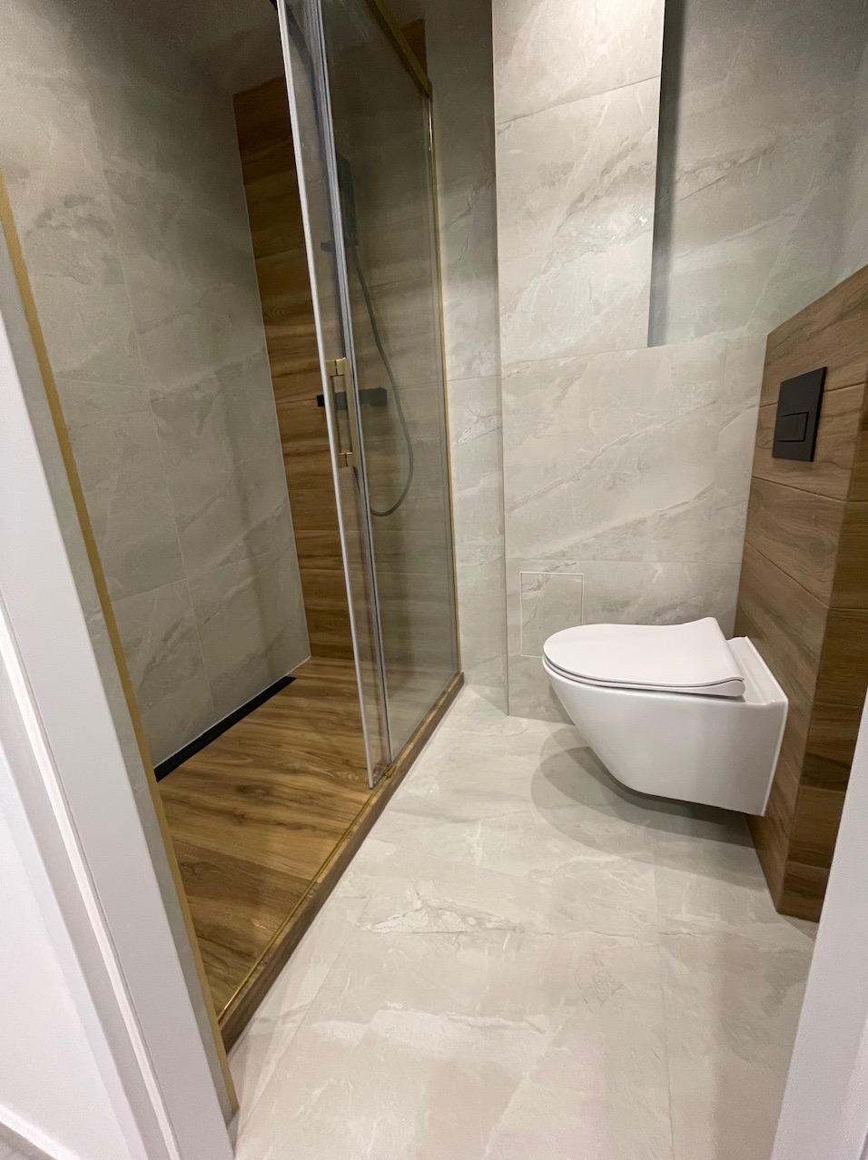 Nowoczesna łazienka z kabiną prysznicową ze złotym obramowaniem, podwieszaną toaletą i marmurowymi płytkami w odcieniach szarości oraz imitacją drewna na podłodze w kabinie.