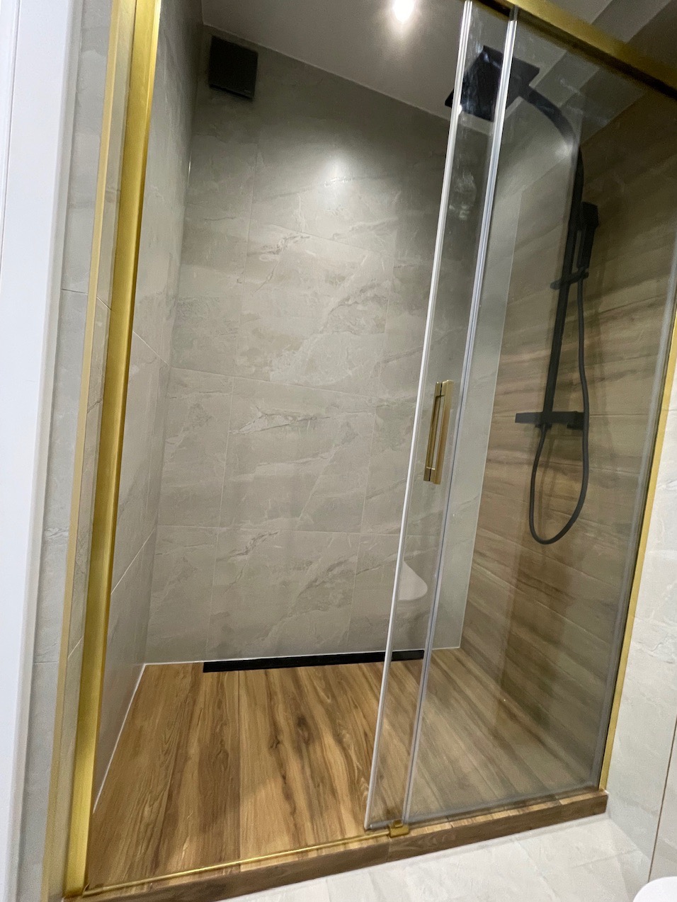 Kabina prysznicowa z przesuwnymi drzwiami, złote ramy, drewnopodobna podłoga, czarna armatura, szare płytki na ścianach.