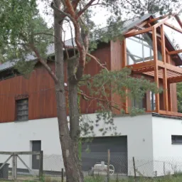 Dwukondygnacyjny dom z drewnianą elewacją i elementami konstrukcyjnymi z drewna, widoczny garaż i ogrodzenie, otoczony drzewami.