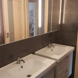 Dwie umywalki z szafkami w szarej łazience z ciemnymi płytkami, oświetlenie ledowe nad lustrem, widok na drzwi do innego pomieszczenia w tle.