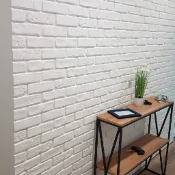 Ściana wyłożona białymi cegłami dekoracyjnymi z widoczną fakturą, uzupełniona konsolą z drewnianymi półkami i metalową konstrukcją, na której stoją ramka na zdjęcie i roślina doniczkowa.