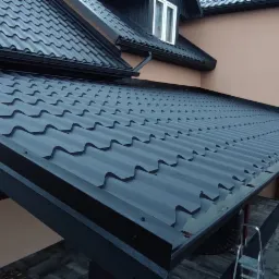 Nowy, grafitowy dach z blachodachówki modułowej, widoczny z boku, z zamontowanymi rynnami i oknem dachowym, na tle beżowej elewacji domu.