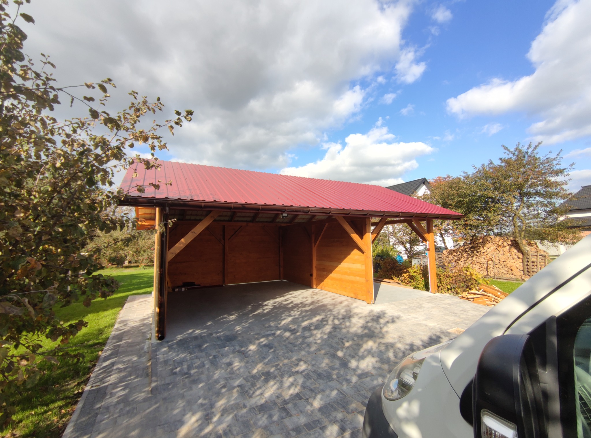 Drewniana wiata garażowa z czerwonym dachem blaszany, kostka brukowa prowadząca do wjazdu, widoczny fragment białego samochodu dostawczego, drzewa w tle.