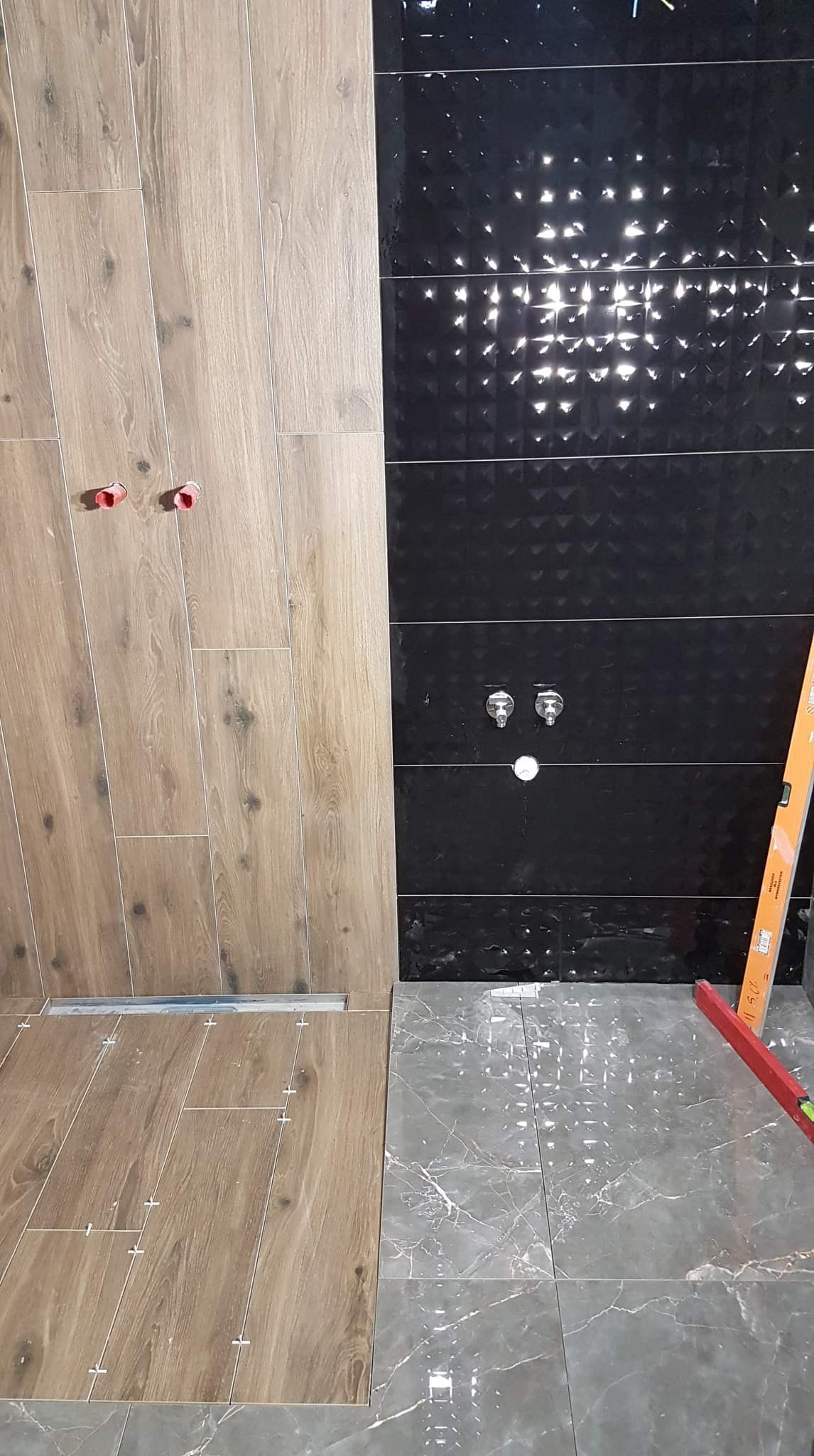 Układanie płytek imitujących drewno na ścianie i podłodze w łazience, widoczne krzyżyki dystansowe i poziomica, obok czarne, błyszczące płytki z geometrycznym wzorem.