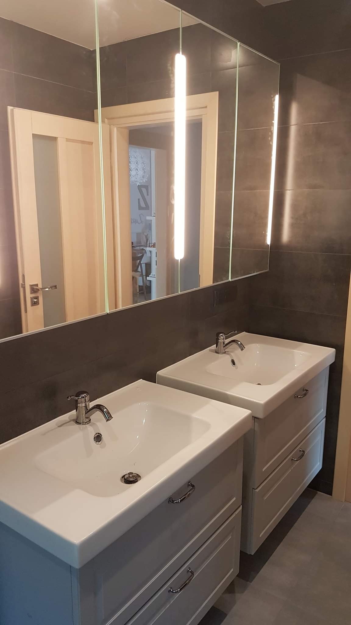 Dwie umywalki z szafkami w szarej łazience z ciemnymi płytkami, oświetlenie ledowe nad lustrem, widok na drzwi do innego pomieszczenia w tle.