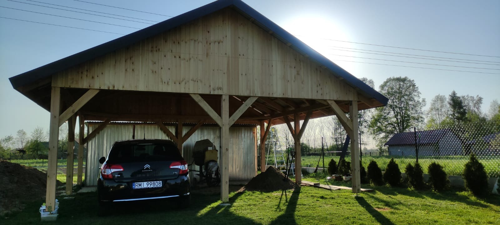 Drewniana wiata garażowa w trakcie budowy, widoczny samochód osobowy, betoniarka i sterta ziemi na trawniku.