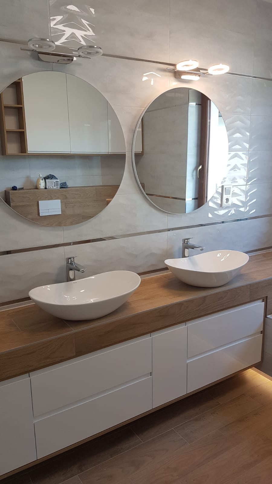 Nowoczesna łazienka z dwoma okrągłymi lustrami, podwieszaną szafką z drewnianym blatem i dwiema asymetrycznymi umywalkami nablatowymi oraz oświetleniem LED