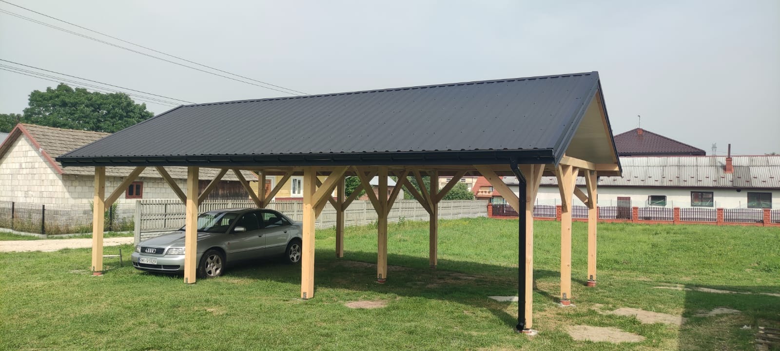 Zadaszenie dla samochodu z drewnianą konstrukcją i czarnym, metalowym dachem na tle wiejskiej zabudowy.