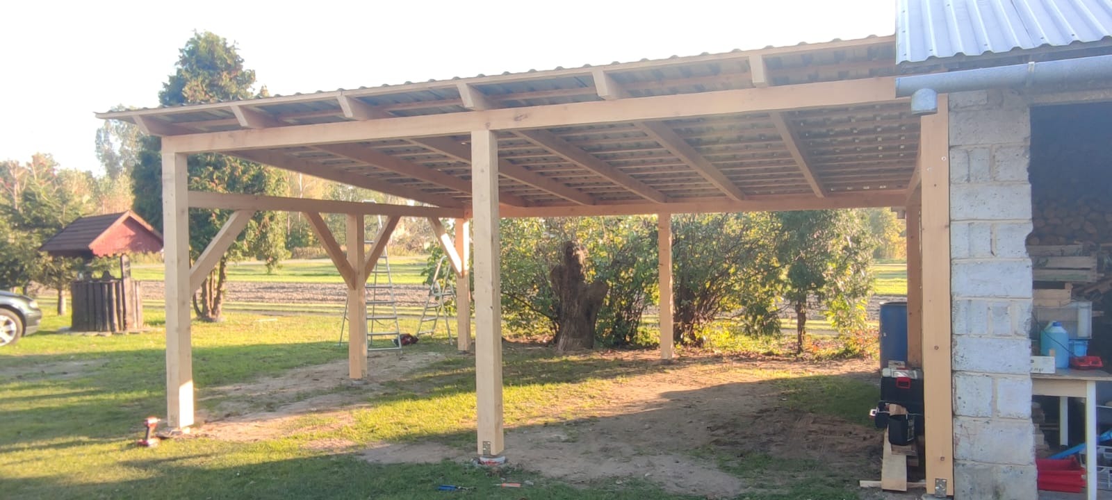 Konstrukcja drewnianej wiaty na podwórku w trakcie budowy, widoczne narzędzia i drabina, w tle studnia i drzewa.