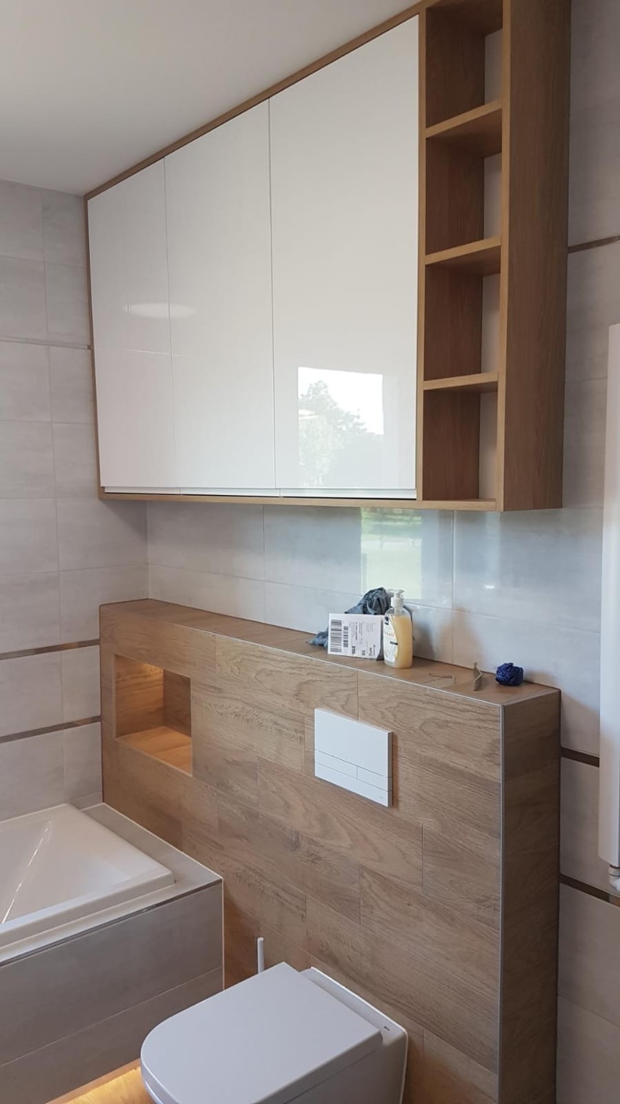 Nowoczesna łazienka z zabudową nad toaletą i wanną, białe, lakierowane szafki wiszące z drewnianymi półkami po prawej stronie, płytki imitujące drewno na ścianach i obudowie wanny, podświetlenie...