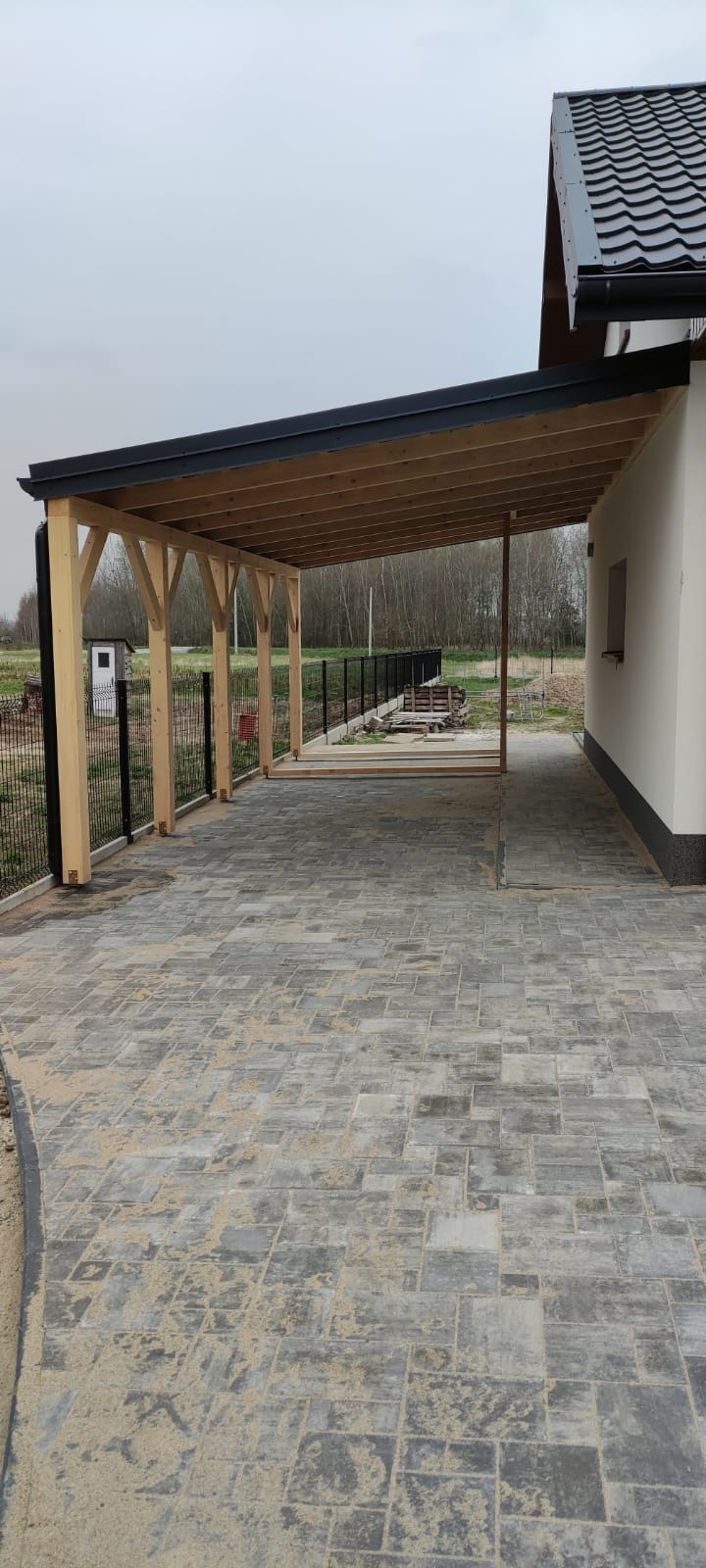 Zadaszenie tarasu z drewnianą konstrukcją wspartą na słupach, wyłożone szarą kostką brukową, widoczne z zewnątrz, z elementami domu w tle.