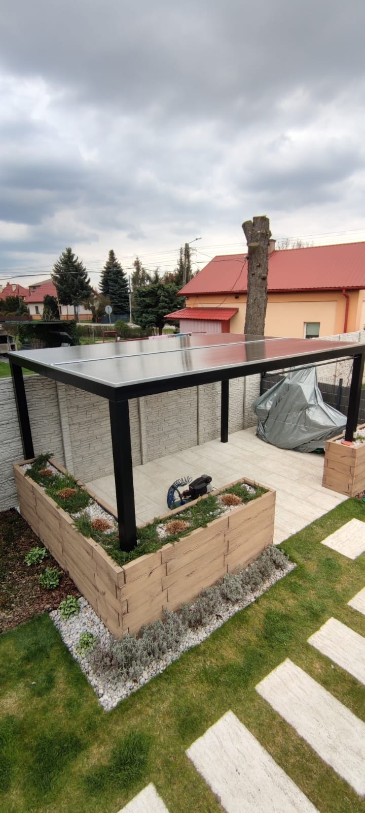 Nowoczesna pergola z czarną konstrukcją i przezroczystym zadaszeniem, obok drewniana donica z roślinami i trawnik z płytami chodnikowymi, w tle dom z czerwoną dachówką.