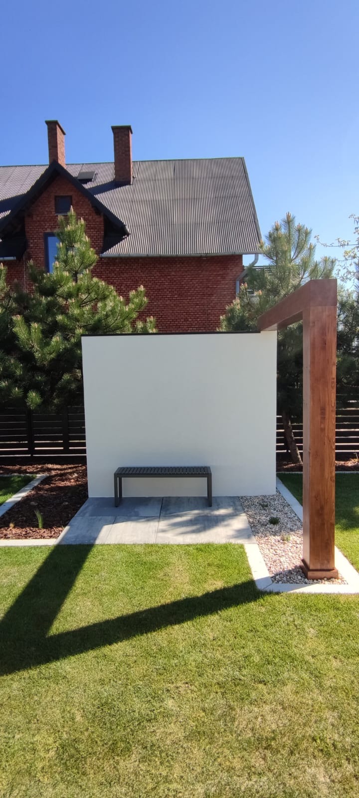 Nowoczesny ogród z minimalistyczną, białą ścianą, drewnianą pergolą i metalową ławką na tle domu z czerwonej cegły i zieleni.