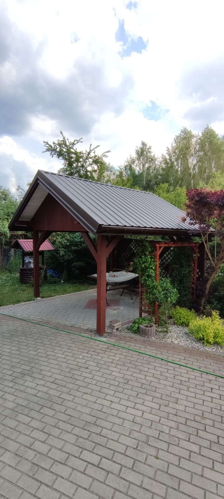Drewniana altana z metalowym dachem i stołem w ogrodzie, widok z zewnątrz na kostkę brukową i zieleń.