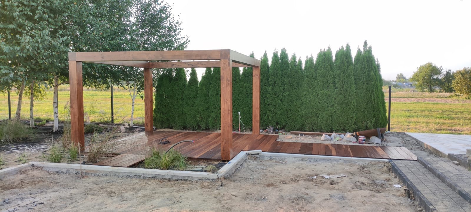 Montaż drewnianej pergoli z tarasem na posesji, widoczne narzędzia i materiały budowlane, tło z tujami i brzozami.