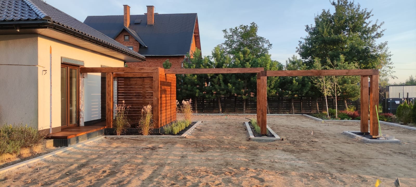 Drewniana pergola z elementami ażurowymi, otoczona świeżo posadzoną roślinnością, na tle domu z czerwonej cegły i piaszczystego podłoża w słoneczny dzień.