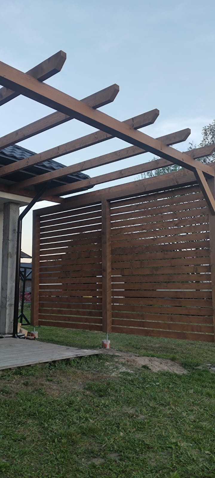 Drewniana pergola z poziomymi listwami, świeżo po montażu, widoczne betonowe podstawy słupków, trawa wokół.