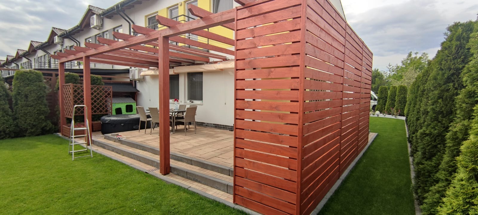 Zadaszony taras z drewnianą pergolą i osłoną boczną, na trawniku drabina, w tle dom jednorodzinny i tuje.