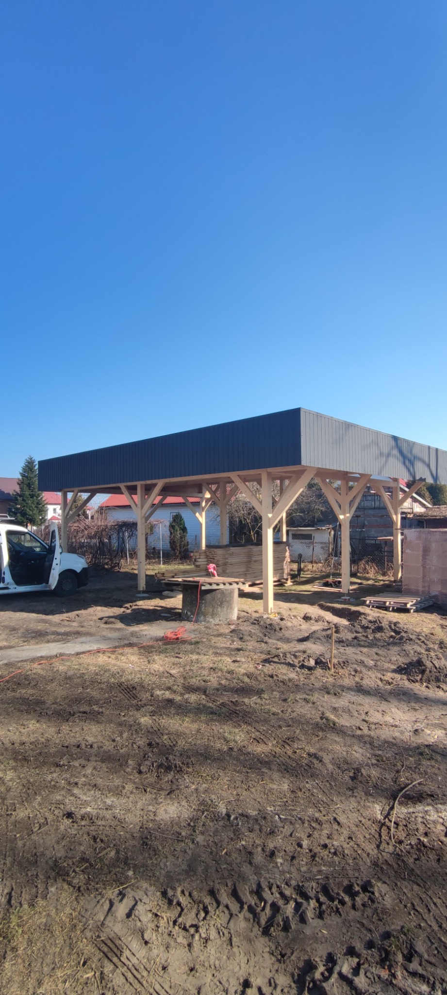 Konstrukcja z drewna i szarej blachy tworząca zadaszenie na zewnątrz, widoczny samochód dostawczy z otwartymi drzwiami, teren w trakcie prac ziemnych.