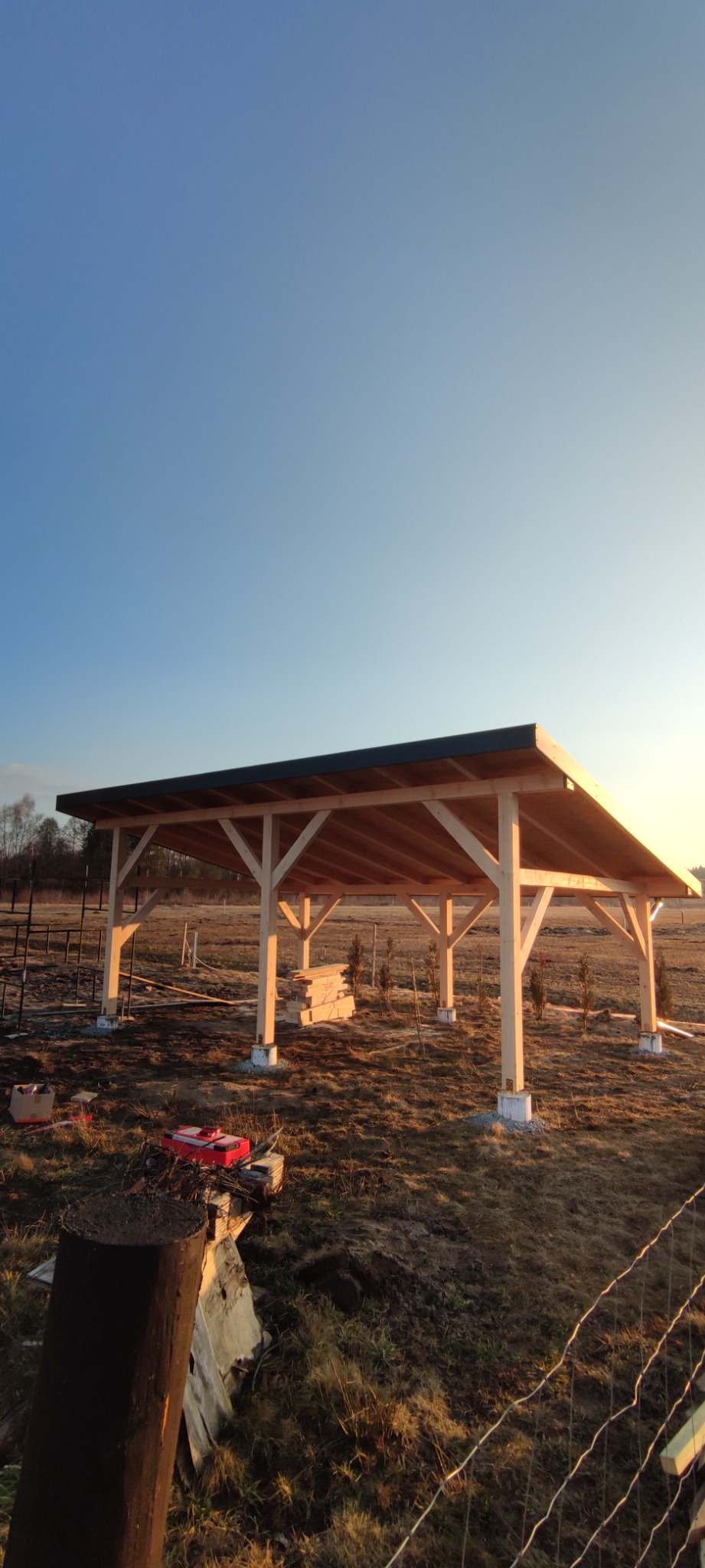 Drewniana konstrukcja z czarnym dachem w trakcie budowy na zewnątrz, widoczne narzędzia i materiały budowlane, niebo w tle.