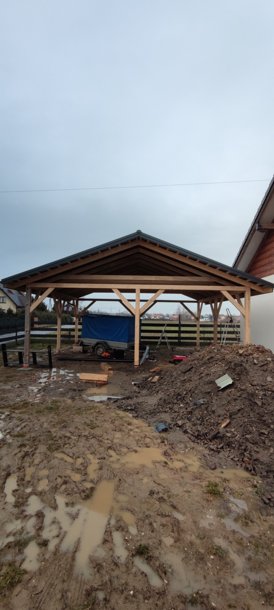 Wznoszenie drewnianej konstrukcji zadaszenia w trakcie budowy, widoczna przyczepka z plandeką i sterta ziemi na błotnistym placu budowy w pochmurny dzień.