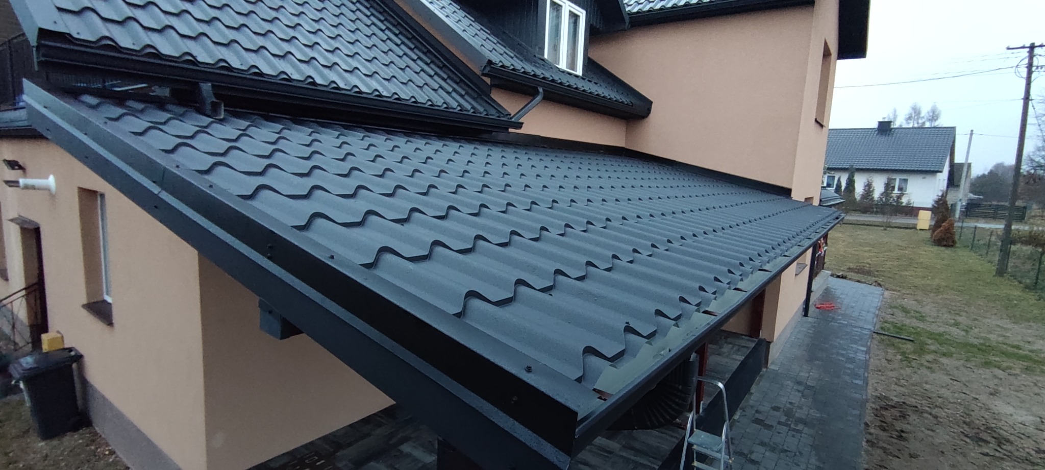 Nowy, grafitowy dach z blachodachówki modułowej, widoczny z boku, z zamontowanymi rynnami i oknem dachowym, na tle beżowej elewacji domu.