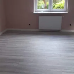 Pomalowane na delikatny różowo ściany pokoju w Nowym Sączu z nową, jasnoszarą podłogą z paneli, białym grzejnikiem pod oknem i widokiem na zieleń za oknem.