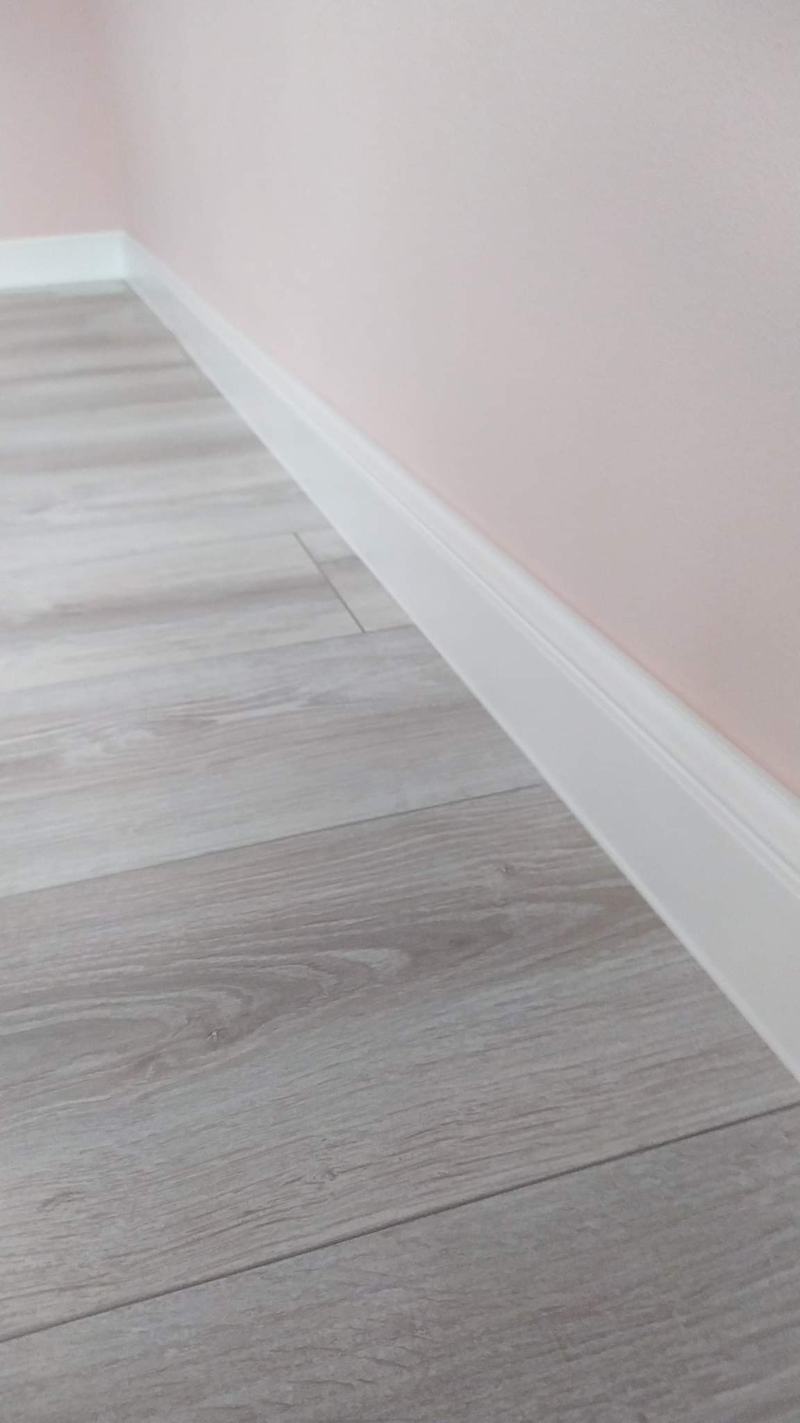 Jasnoszara podłoga panelowa z białą listwą przypodłogową przy różowej ścianie, widok z niskiej perspektywy.