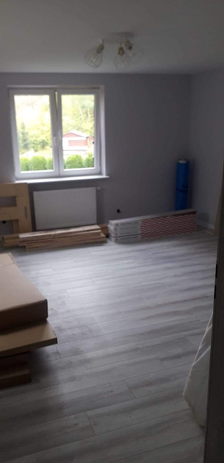 Pokój po remoncie z ułożoną szarą podłogą laminowaną, widoczne materiały do wykończenia, grzejnik, okno z widokiem na zieleń i lampa sufitowa z trzema żarówkami.