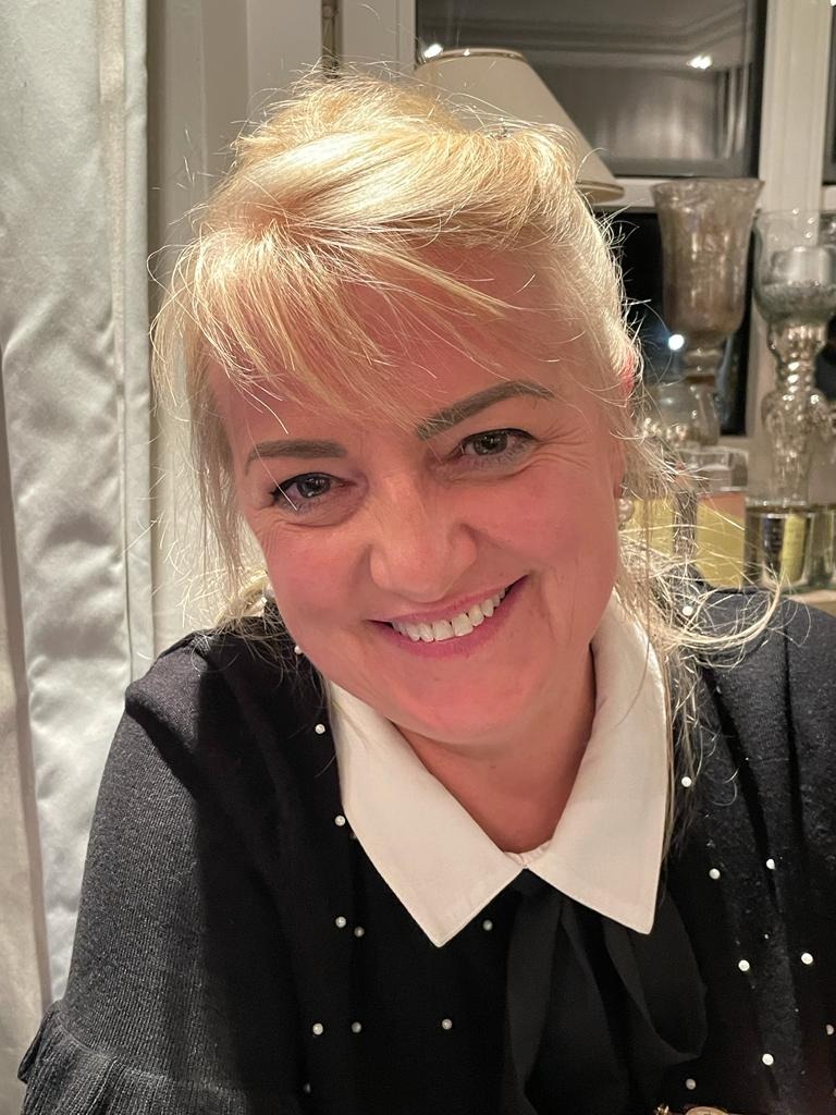 Portret uśmiechniętej kobiety z blond włosami w czarnej bluzce z białym kołnierzykiem i perłami, w tle widoczna lampa i szklane ozdoby.