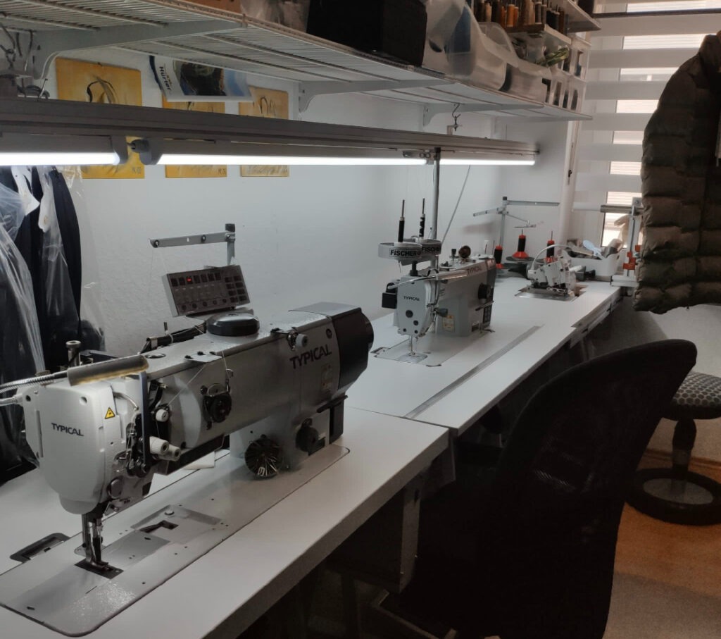 Szwalnia: Rząd nowoczesnych maszyn do szycia marki Typical na białych stołach pod oświetleniem LED, widoczne akcesoria krawieckie na półkach.