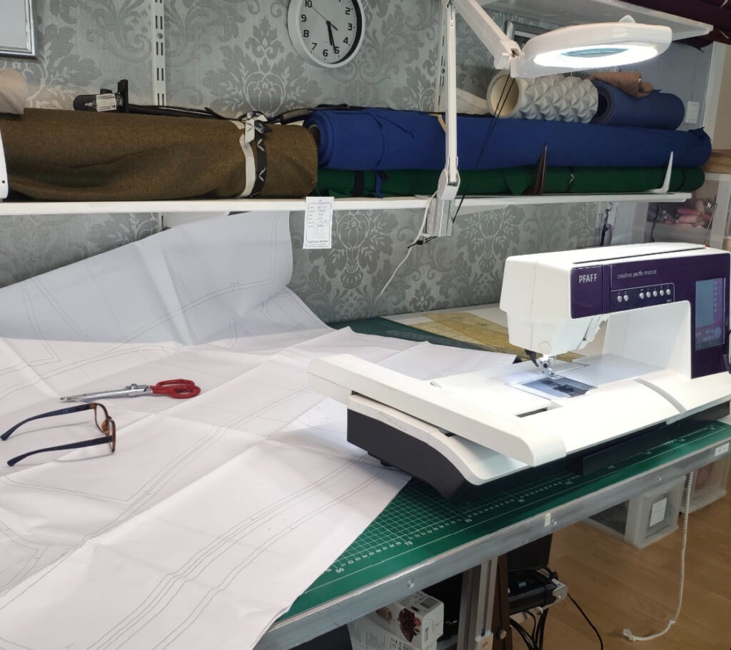 Stół krawiecki z maszyną do szycia Pfaff, rozłożonym szablonem, nożyczkami, okularami i rolkami materiałów na półce w tle.