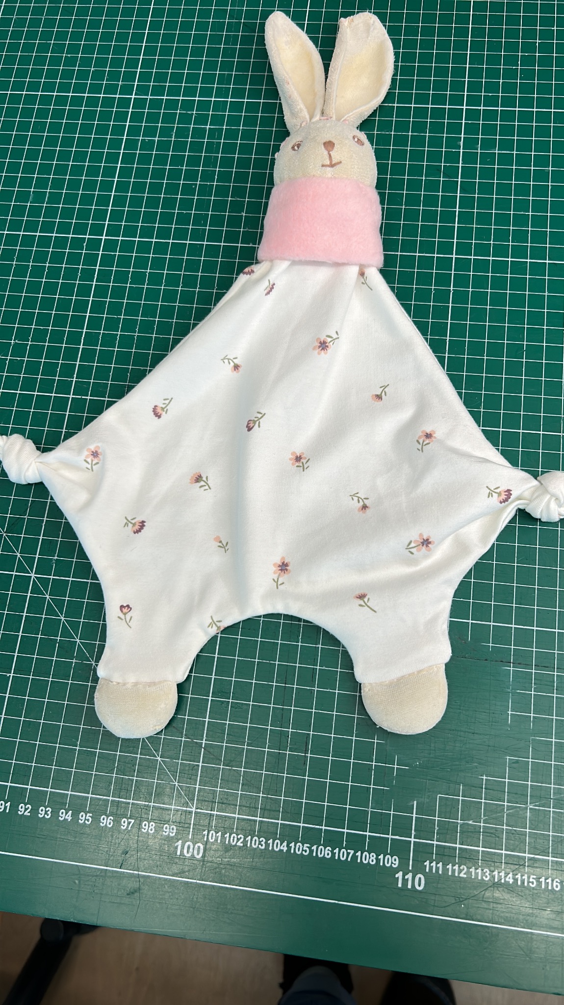 Nierozpoznany, biały kocyk-przytulanka w kształcie rombu z głową królika, różowym szalikiem i drobnymi kwiatkami, leżący na zielonej macie do cięcia z podziałką centymetrową.