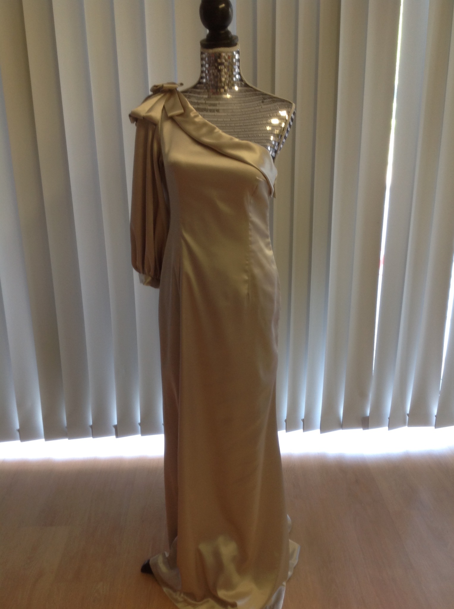 Satynowa suknia wieczorowa w kolorze szampana na manekinie z cekinową górą, z drapowanym ramieniem i pojedynczym długim rękawem, na tle białych żaluzji.