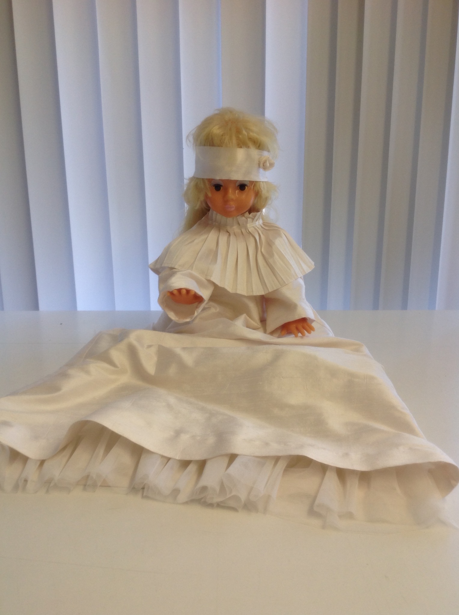 Lalka w białej sukience z plisowanym kołnierzem i opaską, siedząca na białym stole na tle białych żaluzji.