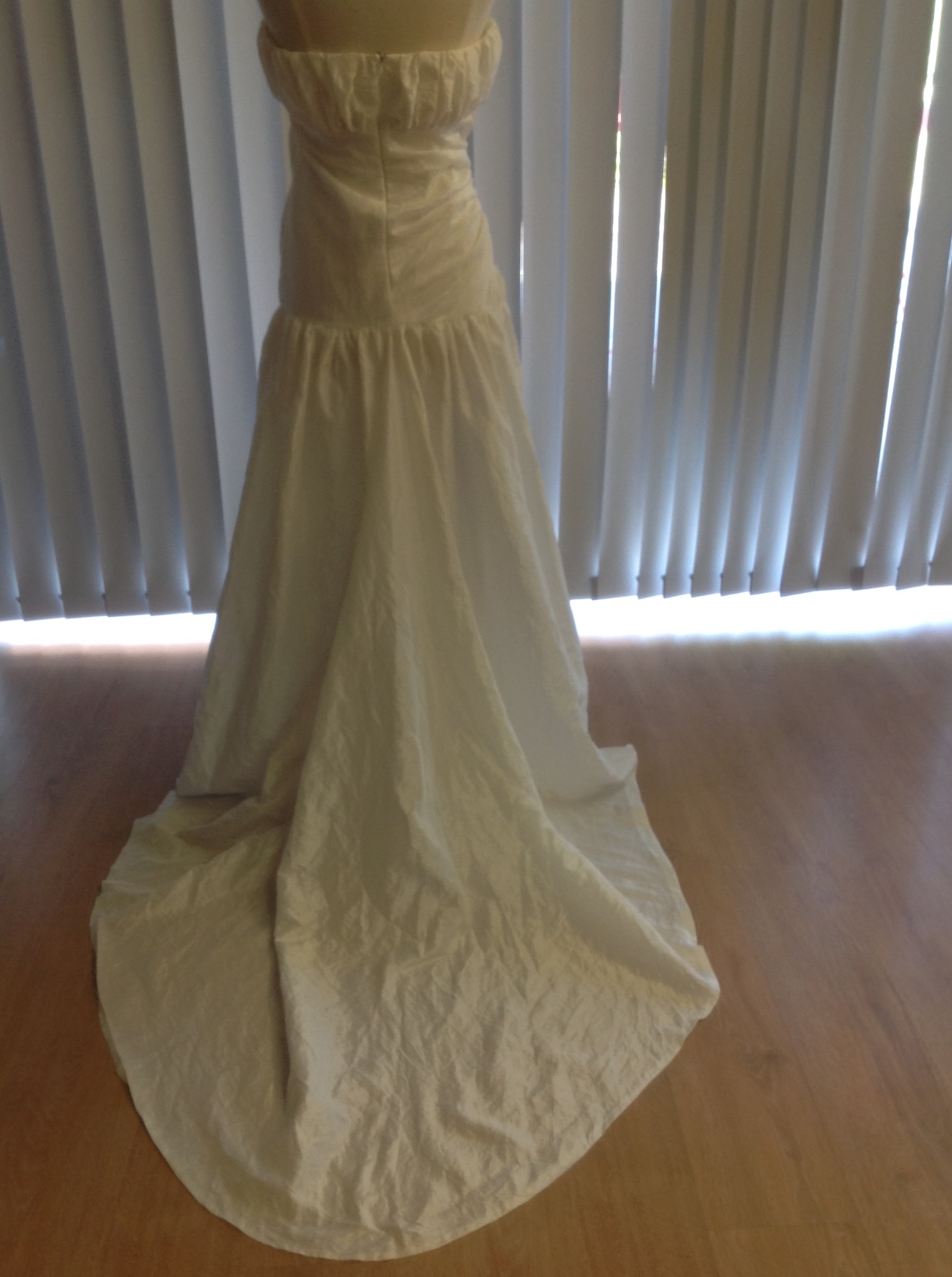 Widok z tyłu białej sukni bez ramiączek na manekinie, z marszczeniami w talii i długim, lekko pogniecionym dołem, na tle okna z żaluzjami.