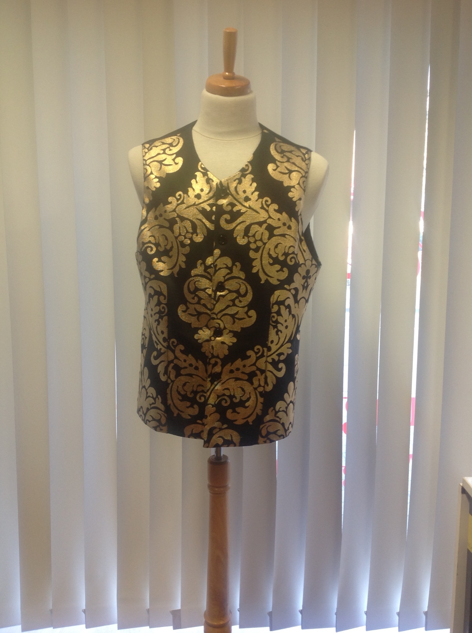 Czarna kamizelka z wytłaczanym złotym wzorem na drewnianym manekinie, na tle białych żaluzji.