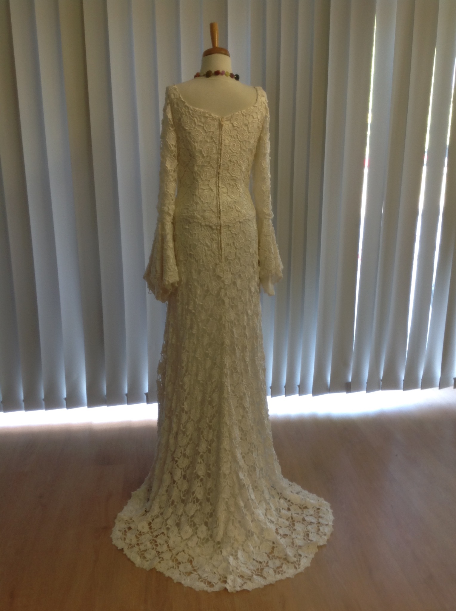 Długa suknia ślubna z koronki, eksponowana na manekinie z drewnianą szyją i kolorowym naszyjnikiem, widziana od tyłu na tle białych żaluzji.