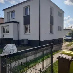 Dwupiętrowy dom z jasną elewacją i ciemnymi akcentami wokół okien oraz u podstawy, widziany zza metalowego ogrodzenia, z workiem materiałów budowlanych i koszami na śmieci w pobliżu.