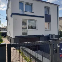 Dwupiętrowy dom z ocieploną elewacją w odcieniach szarości i bieli, widoczny zza czarnego ogrodzenia panelowego, z numerem '14' na ścianie.