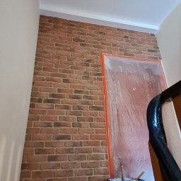 Kleczka Adam Usługi Remontowo - Wykończeniowe - Klatka schodowa w trakcie zabezpieczania przed malowaniem, ściana z cegły dekoracyjnej, zabezpieczone folią drzwi, wiadro z farbą na schodach.