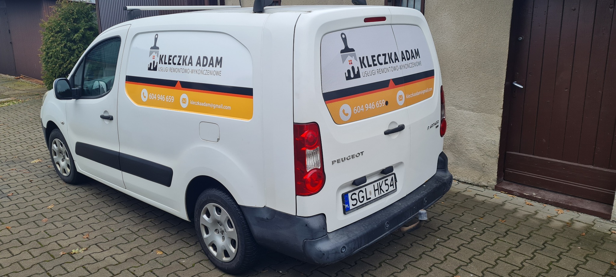 Biały samochód dostawczy firmy remontowo-wykończeniowej 'KLECZKA ADAM' z numerem telefonu i adresem e-mail na boku i tylnej szybie, zaparkowany na kostce brukowej.