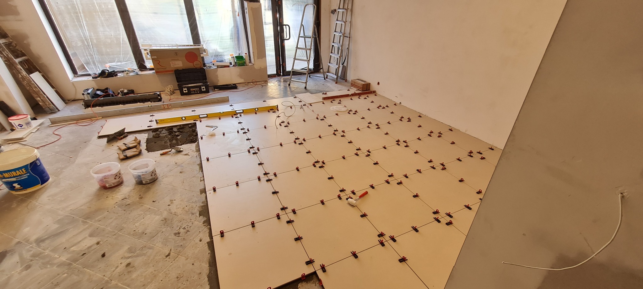 Układanie beżowych płytek podłogowych w salonie jubilerskim, widoczne czerwone klipsy poziomujące, poziomica, wiadro z klejem, narzędzia i drabina w tle.