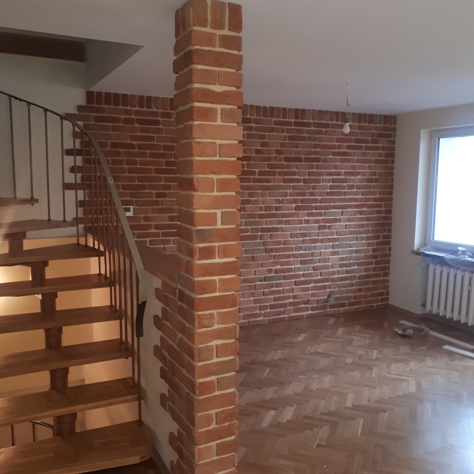 Ściana wykończona ciętą cegłą w odcieniach czerwieni i brązu, z widoczną kolumną również obłożoną cegłą, drewniane schody spiralne z metalową balustradą oraz parkiet w jodełkę na podłodze.