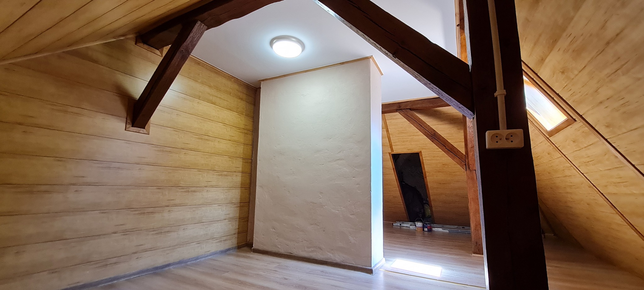 Wykończone poddasze z drewnianą boazerią, białą ścianką działową i widocznymi elementami konstrukcji drewnianej, oświetlone centralną lampą sufitową i światłem z okna dachowego.